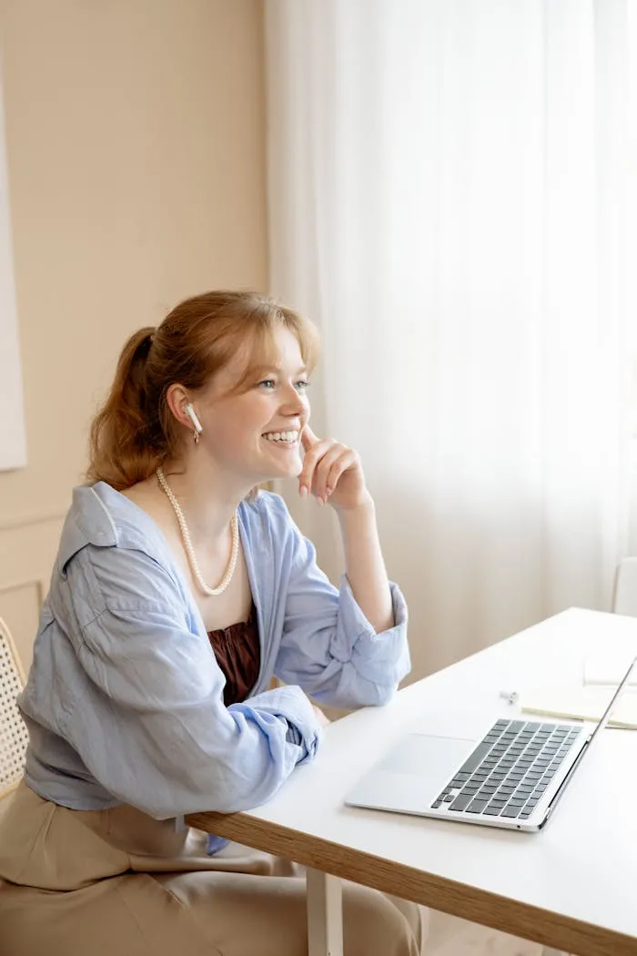 A smiling businesswoman with earbuds working on her laptop at a wooden desk in a bright office.