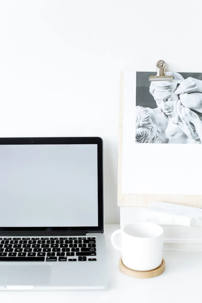 Elegant minimalist workspace featuring a closed laptop, coffee mug, and artistic clipboard in a bright setting.